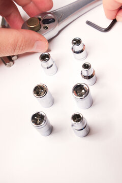 Close‑up of a mechanical tool arrangement on a white surface. A ratchet wrench with a gold directional switch lies near seven chrome sockets of varying sizes, a small L‑shaped Allen key