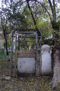 An old, weathered metal gate and a white concrete pillar stand framed by autumn trees with fallen leaves on the ground in Osh, Kyrgyzstan