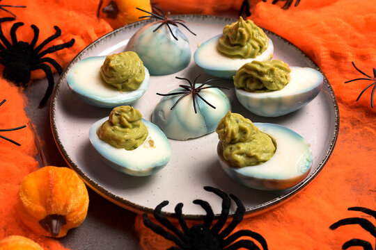 Halloween deviled eggs stuffed with avocado guacamole cream on a plate decorated with black spiders and orange cobwebs for spooky holiday party food
