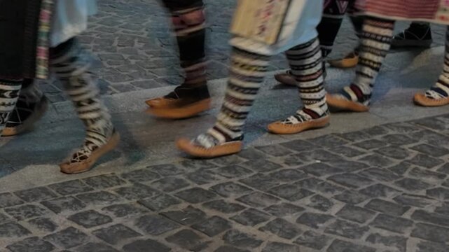People wearing traditional patterned socks and leather shoes walk across a dark cobblestone street. Such lively cultural parades celebrate vibrant historical folklore and community heritage.