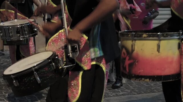 Musicians in sparkling costumes play drums energetically on a dark cobblestone street. The vibrant rhythmic performance celebrates cultural heritage and brings joyful energy to the night.