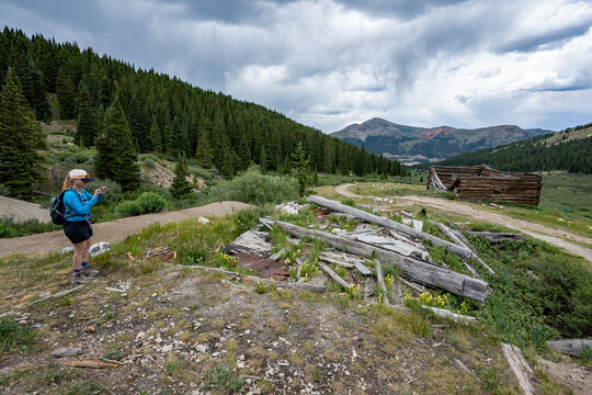Woman hiker exploring abandoned mining cabins on Mayflower Gulch trail near Copper Mountain, Colorado under summer afternoon cloudscape.