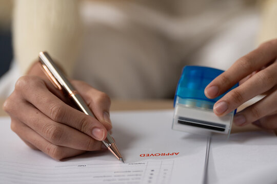 Document approval concept with a person signing paperwork beside an approved stamp at office desk, showing approval, document, signature, authorization, verification
