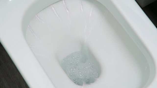 White toilet bowl drains water in flushing sequence. Water swirls and bubbles as it empties. Clean porcelain gleams under soft lighting. Modern bathroom design shown in detail. Use for home improvemen