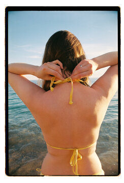 A woman ties her yellow bikini at the water's edge