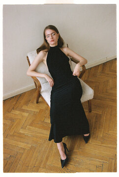 Model poses in a black dress with high heels sitting on a chair