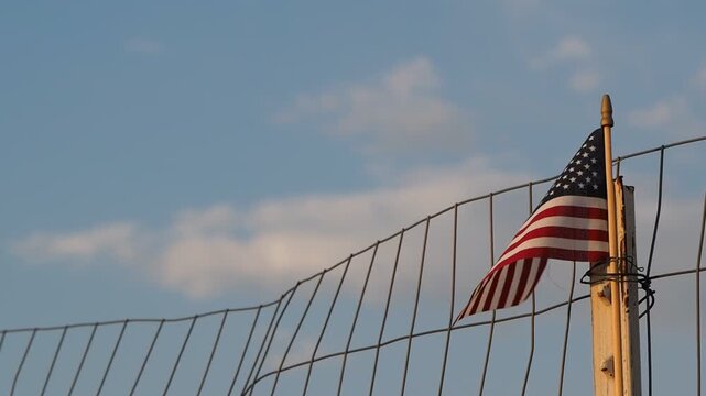 A small, late afternoon lit American flag blowing in the wind with a partly cloudy blue sky in the background held on to a post with wire holding up a fence. Eye level.