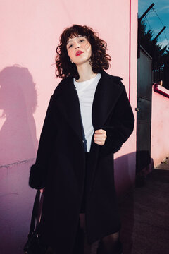 Young woman posing with black coat by pink wall