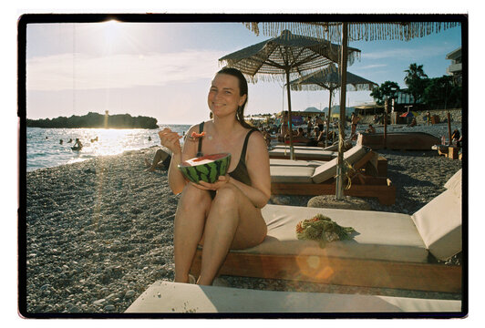 A woman eats watermelon on a pebble beach at sunset