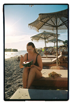 A woman eats watermelon on a pebble beach at golden hour