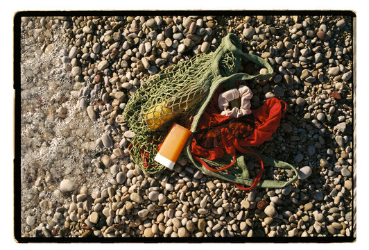 Green mesh bag with sunscreen and a red bikini on a pebble beach
