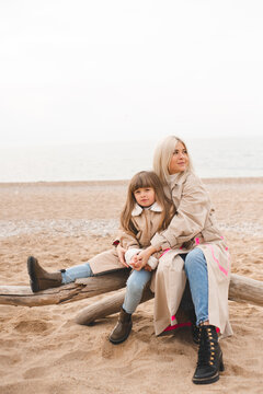 Mother with child daughter sitting on log at sandy beach wearing similar autumn beige trench coat over sea. Motherhood.