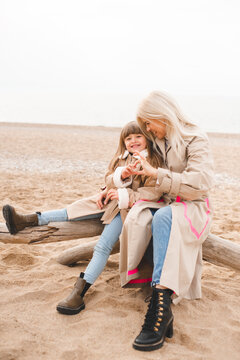 Mother with child daughter sitting on log at sandy beach wearing similar autumn beige trench coat over sea. Motherhood.