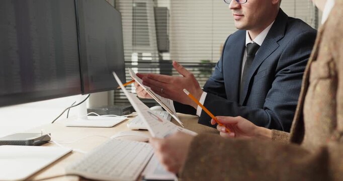 Colleagues perform finance analysis reviewing charts. Two professionals compare printed graphs and discuss a report beside a computer monitor at a clean desk. Clear concept of team finance.
