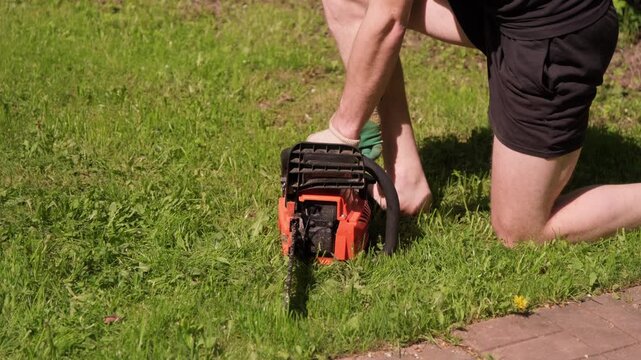 Close-up of a man in shorts starting a chainsaw by pulling the lever during daylight hours