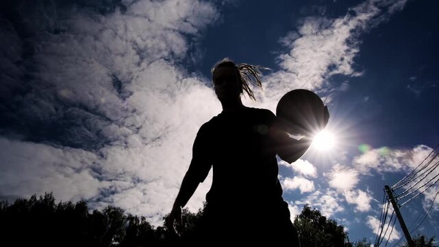 A silhouette of a man with long dreadlocks playing basketball, bouncing the ball on the floor against the backdrop of a bright sky and bright sun