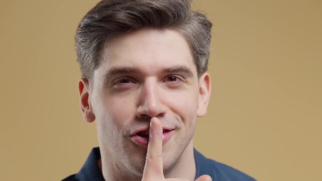 Smiling young man making a shhh gesture with a finger on his lips. Studio portrait on a yellow background. Privacy, confidentiality, secret information and silence.
