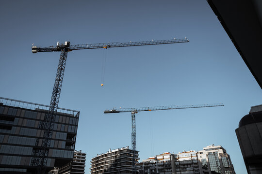 Two tower cranes rise above modern apartment blocks in a low angle urban view, illustrating active construction, real estate growth and city development under clear blue sky