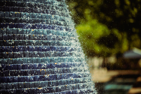 Sparkling water streams over the stepped blue mosaic surface of a conical outdoor fountain, creating lively splashes, droplets and cooling texture against soft park greenery