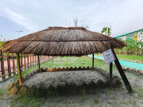 Thatched roof gazebo with clay bricks border and sandy floor in park under cloudy sky with green grass surrounding it.