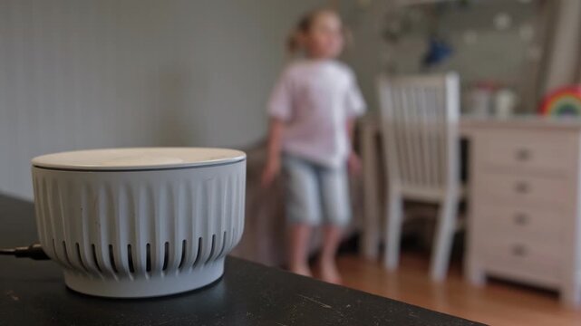 Smart speaker on table playing music for dancing little girl on blurred background. Cute child listening to audio voice assistant in bedroom. Modern smart home, AI technology and lifestyle concept.