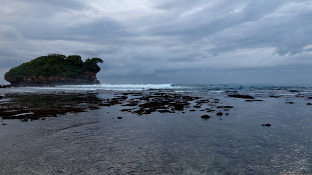 Serene coastal scenery featuring a verdant rocky island, shallow tidal pools, gentle surf, and a dramatic overcast sky along the shoreline in Pacitan, Indonesia