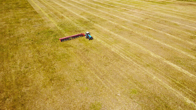 A high-angle aerial photograph capturing a blue tractor pulling a red trailing mower swather across a wide agricultural field