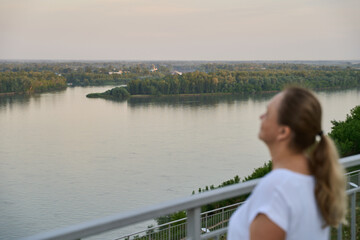 Fototapeta premium Profile Woman Gazing Over River From Terrace At Sunset With Forested Shoreline And Quiet Water Under Pale Evening Sky View.
