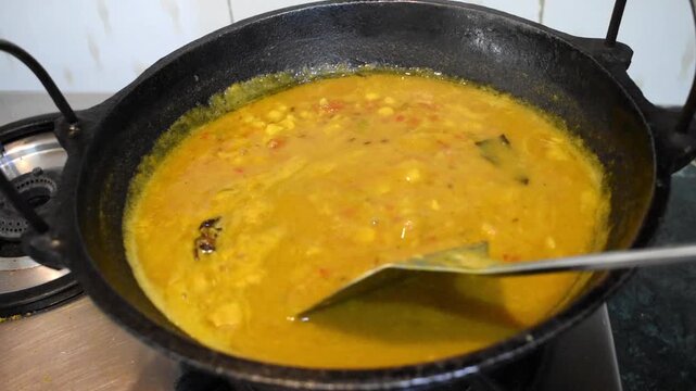 stirring freshly cooked dal with a spoon in a traditional cast iron kadhai in an Indian home kitchen