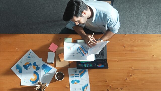 Top aerial view of manager working on laptop while looking at data analysis. Leader compare financial statistic graph while making decision at meeting table with document scatter around. Convocation.