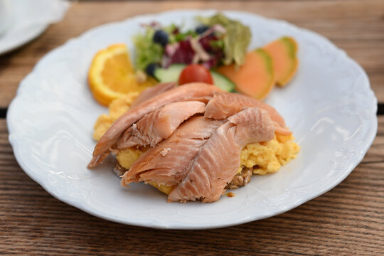 Juicy smoked salmon on scrambled eggs and bread with a colorful salad garnish on a white plate and a rustic wooden table, selected focus