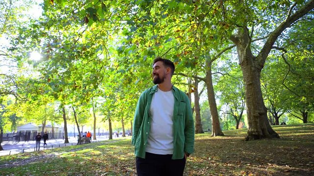 A cheerful adult enjoys a peaceful stroll outdoors under large leafy trees during a bright day. He is smiling and dressed in a casual green shirt and white t-shirt.
