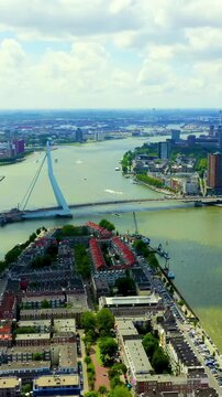 Vertical video. Rotterdam, Netherlands. Erasmus Bridge, its 139-meter asymmetric steel pylon and sweeping cable stays spanning the Nieuwe Maas, Drone footage over Noordereiland under a partly cloudy 