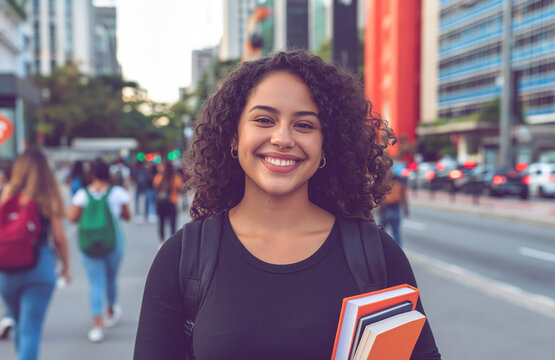 Young smiling student with curly hair holding books on a busy city street