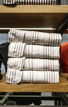 Stack of folded light colored men's shirts with vertical stripes displayed on a wooden shelf, representing classic menswear, fashion retail and clothing organization.