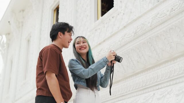 Happy young Asian couple taking selfie video and exploring beautiful white temple architecture. Cheerful romantic tourist lovers enjoying cultural sightseeing summer vacation.