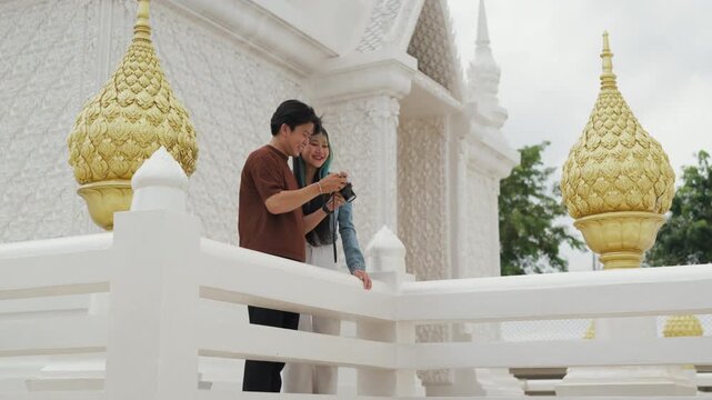Happy young Asian couple taking selfie video and exploring beautiful white temple architecture. Cheerful romantic tourist lovers enjoying cultural sightseeing summer vacation.
