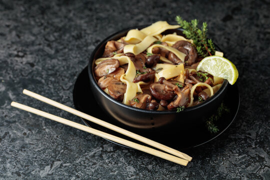 Shimeji and shiitake mushrooms stir-fried with soy sauce, garlic and thyme, served with noodles and lime slice in a black ceramic bowl.