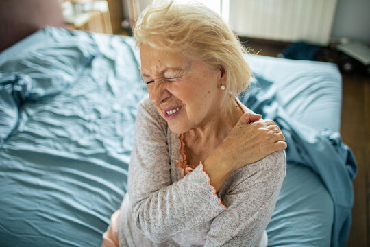 Senior woman grimacing with shoulder pain on bed at home
