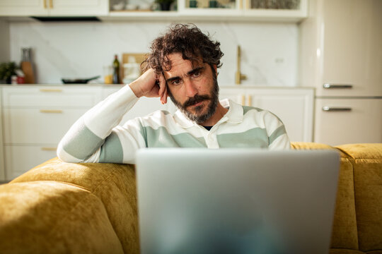 Tired bearded man working on laptop on couch in home kitchen
