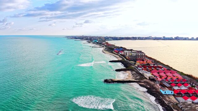 Aerial view of a coastal town along turquoise sea, Bulgaria. Drone perspective of red roofed buildings, breakwaters and cityscape in the distance.