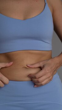 Close-up of young woman pinching soft belly fat and skin fold in blue sportswear, showing abdominal fat, body shape and weight loss concern on gray background
