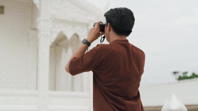 Handsome young Asian male tourist taking beautiful photos with digital camera at white temple. Cheerful solo traveler enjoying cultural sightseeing adventure on summer vacation.