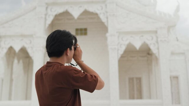 Handsome young Asian male tourist taking beautiful photos with digital camera at white temple. Cheerful solo traveler enjoying cultural sightseeing adventure on summer vacation.