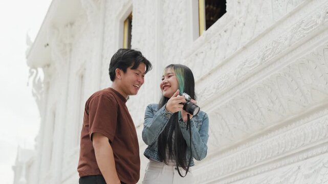 Attractive young Asian couple looking at digital camera pictures at beautiful white temple. Happy romantic tourist travelers enjoying historic sightseeing summer holiday vacation.