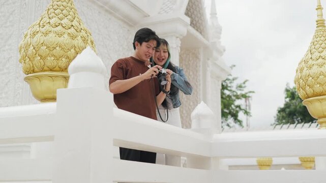 Attractive young Asian couple looking at digital camera pictures at beautiful white temple. Happy romantic tourist travelers enjoying historic sightseeing summer holiday vacation.