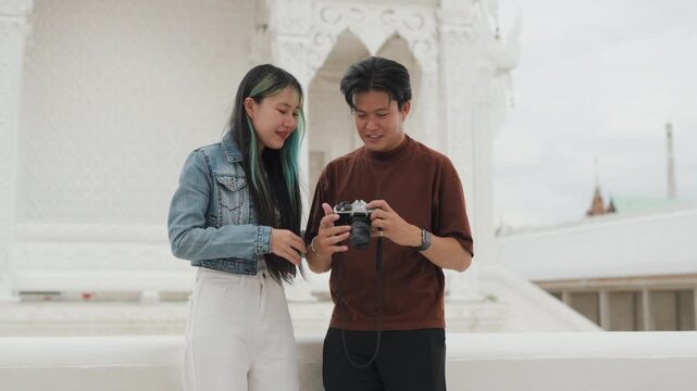 Attractive young Asian couple looking at digital camera pictures at beautiful white temple. Happy romantic tourist travelers enjoying historic sightseeing summer holiday vacation.