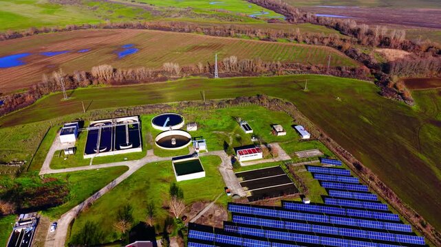 Aerial view of wastewater treatment plant beside solar farm, Bulgaria. Circular tanks, green fields and a winding river fill the drone frame.