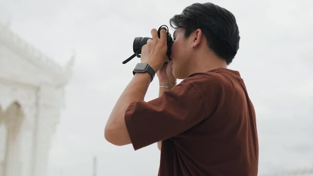 Handsome young Asian male tourist taking beautiful photos with digital camera at white temple. Cheerful solo traveler enjoying cultural sightseeing adventure on summer vacation.