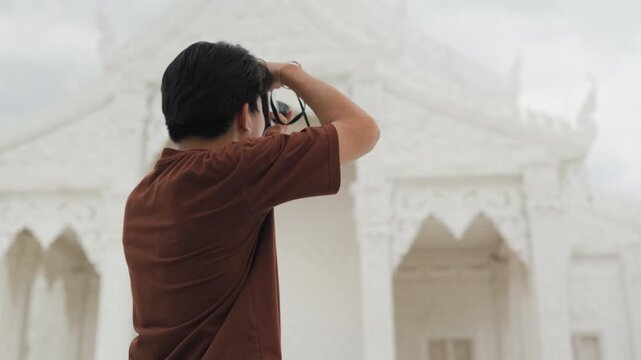 Handsome young Asian male tourist taking beautiful photos with digital camera at white temple. Cheerful solo traveler enjoying cultural sightseeing adventure on summer vacation.